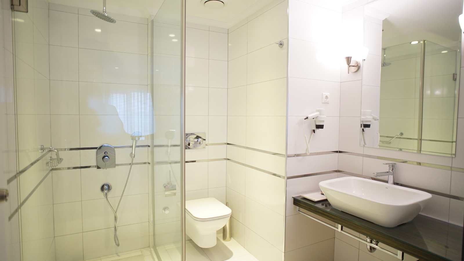 Beyaz fayanslar, cam duşakabin ve modern lavabo detayıyla tasarlanmış otel banyosu.