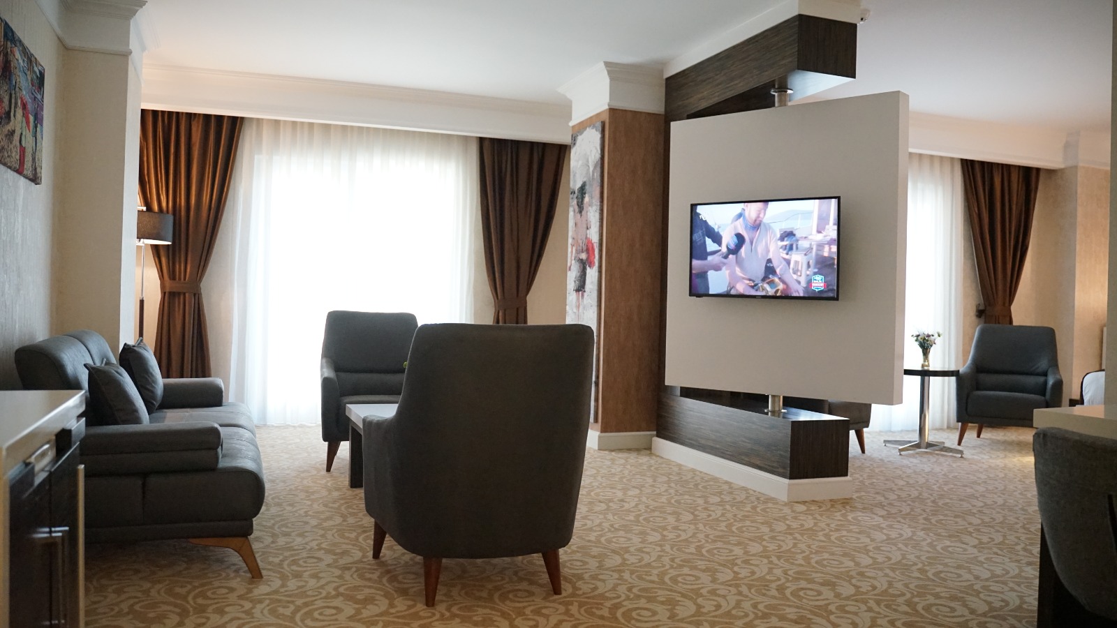 Modern koltuklar, TV ünitesi ve ahşap panellerle tasarlanmış otel dinlenme alanı.