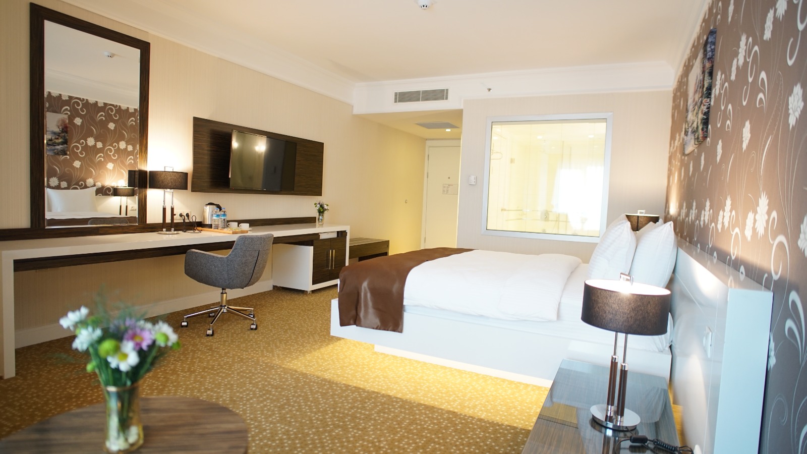 Çalışma masası, konforlu yatak ve modern banyo cam duvarlı otel yatak odası.