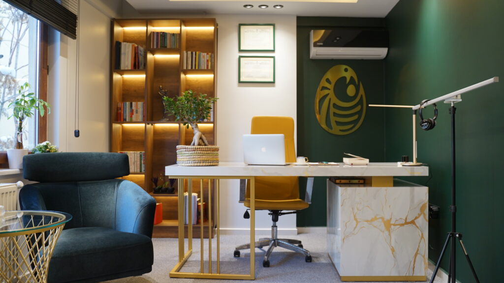 Psikolojik Danışmanlık Ofis Tasarımı Uzman Psikolojik Danışman Gamze Ağbaba'nın, yeşil duvar ve aydınlatmalı kitaplıkla tasarlanmış çalışma masası ve ofisi.